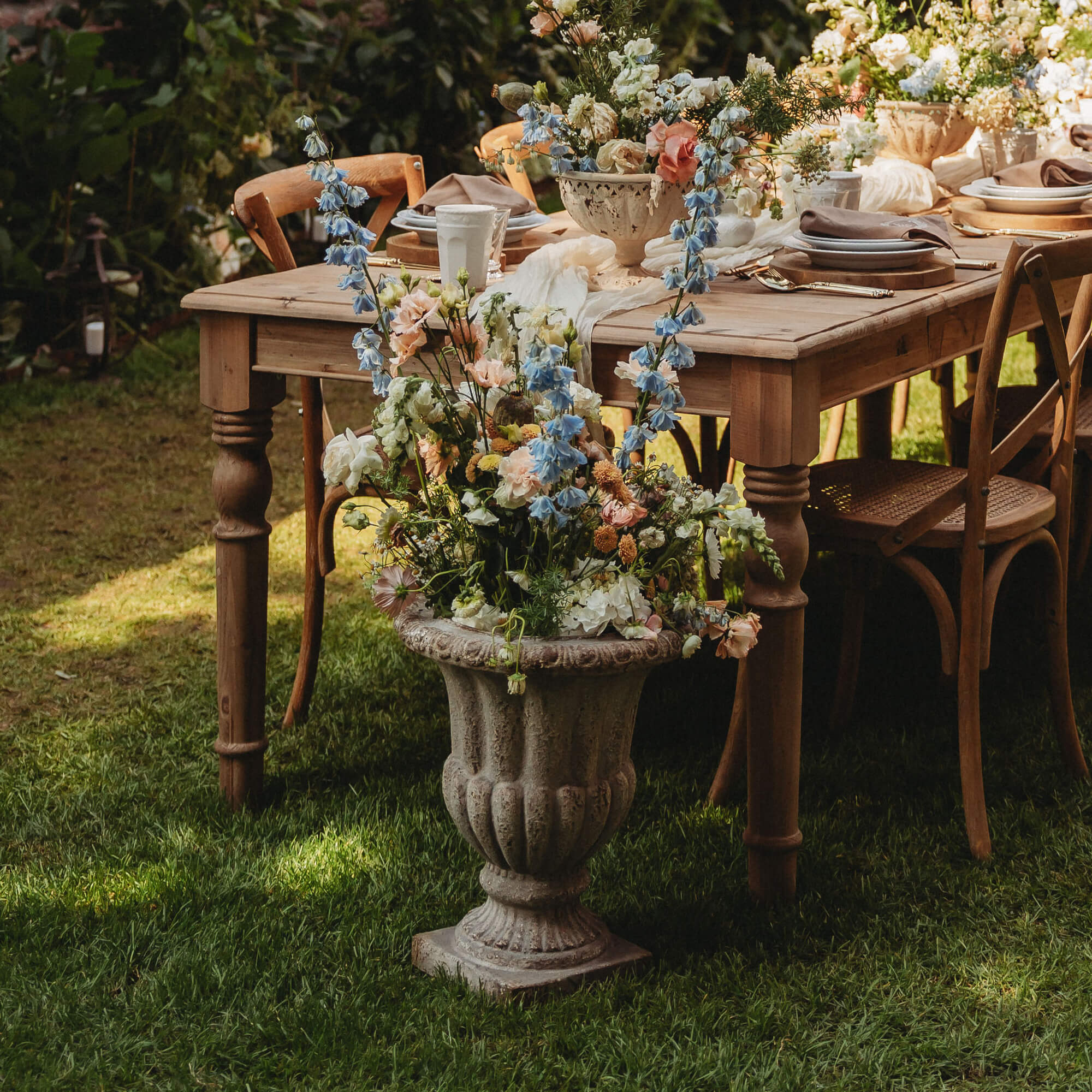 Grand vase en pierre avec fleurs pastel devant une table en bois et chaises dans un jardin verdoyant