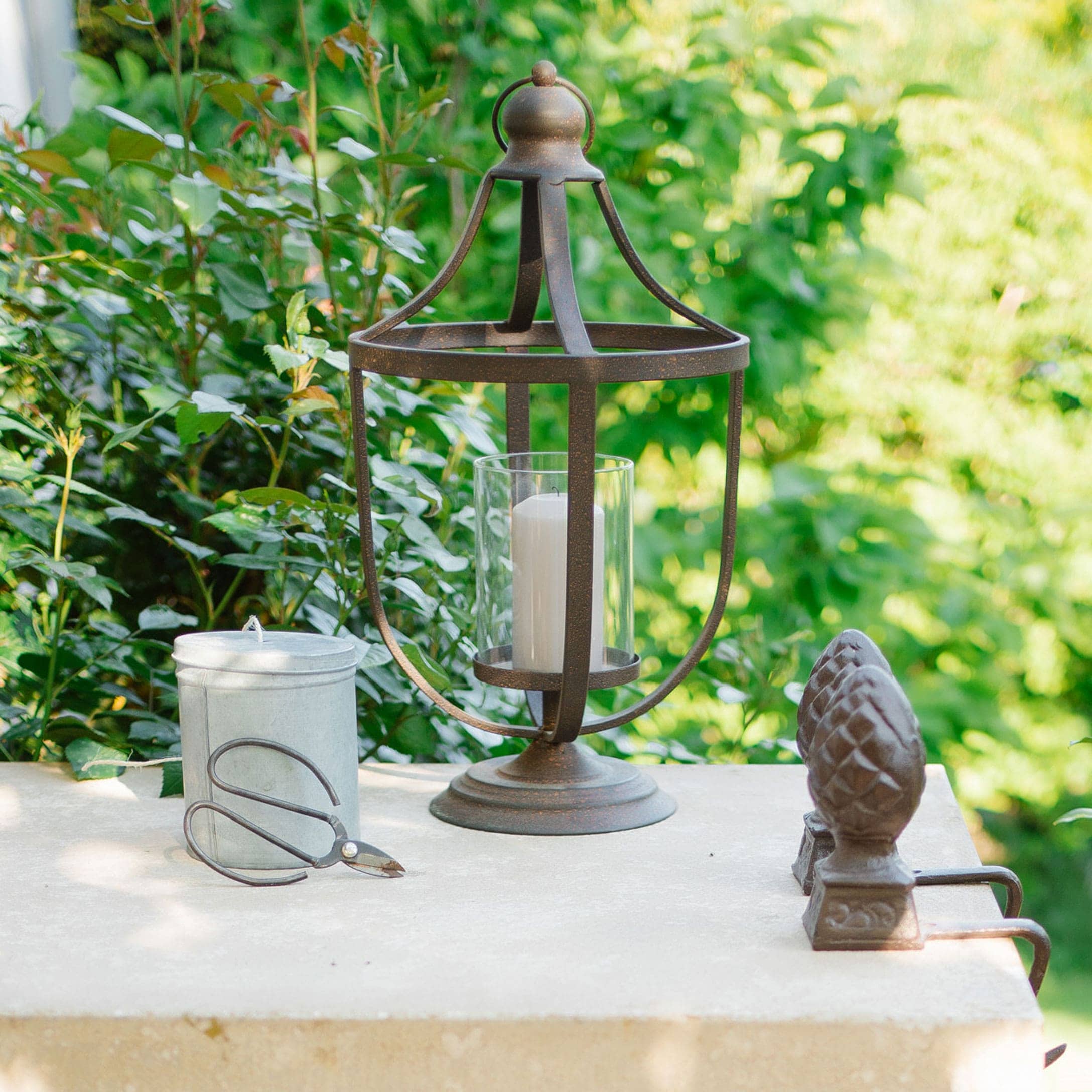 Lanterne en métal avec bougie blanche et outils de jardinage sur table claire devant un feuillage vert