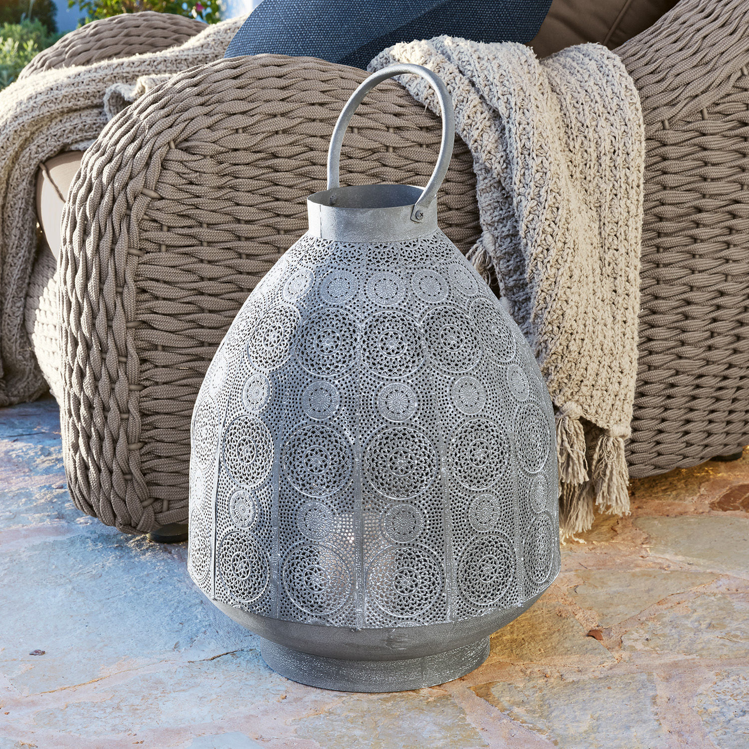 Lanterne Morlet Lanterne en métal gris avec motif ajouré posée sur un sol en pierre près d'un fauteuil en osier et d'un plaid