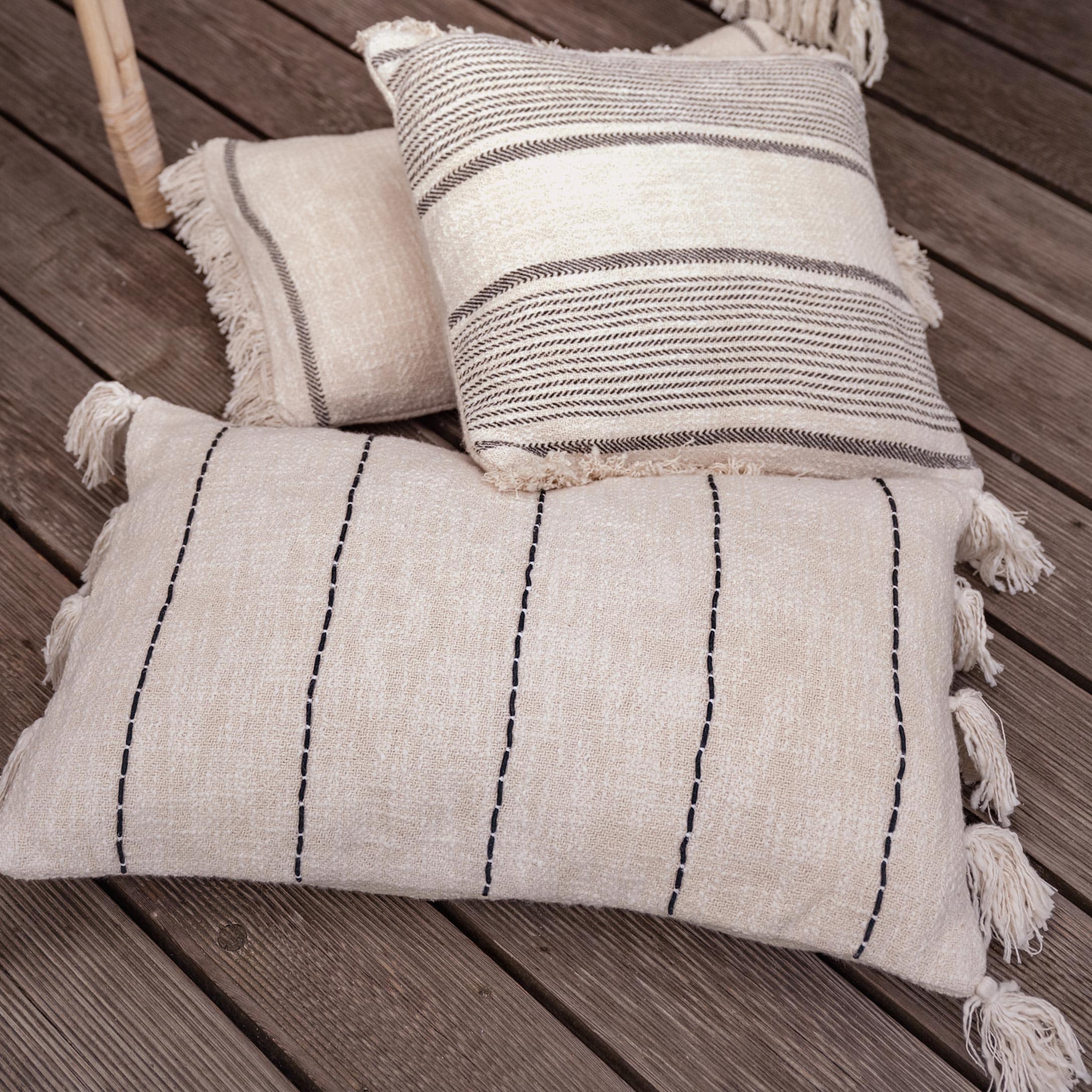 Coussins décoratifs en coton beige avec franges et rayures noires posés sur un sol en bois
