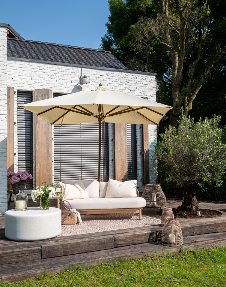 Terrasse avec mur en brique blanche, canapé beige avec coussins et parasol crème sur terrasse en bois
