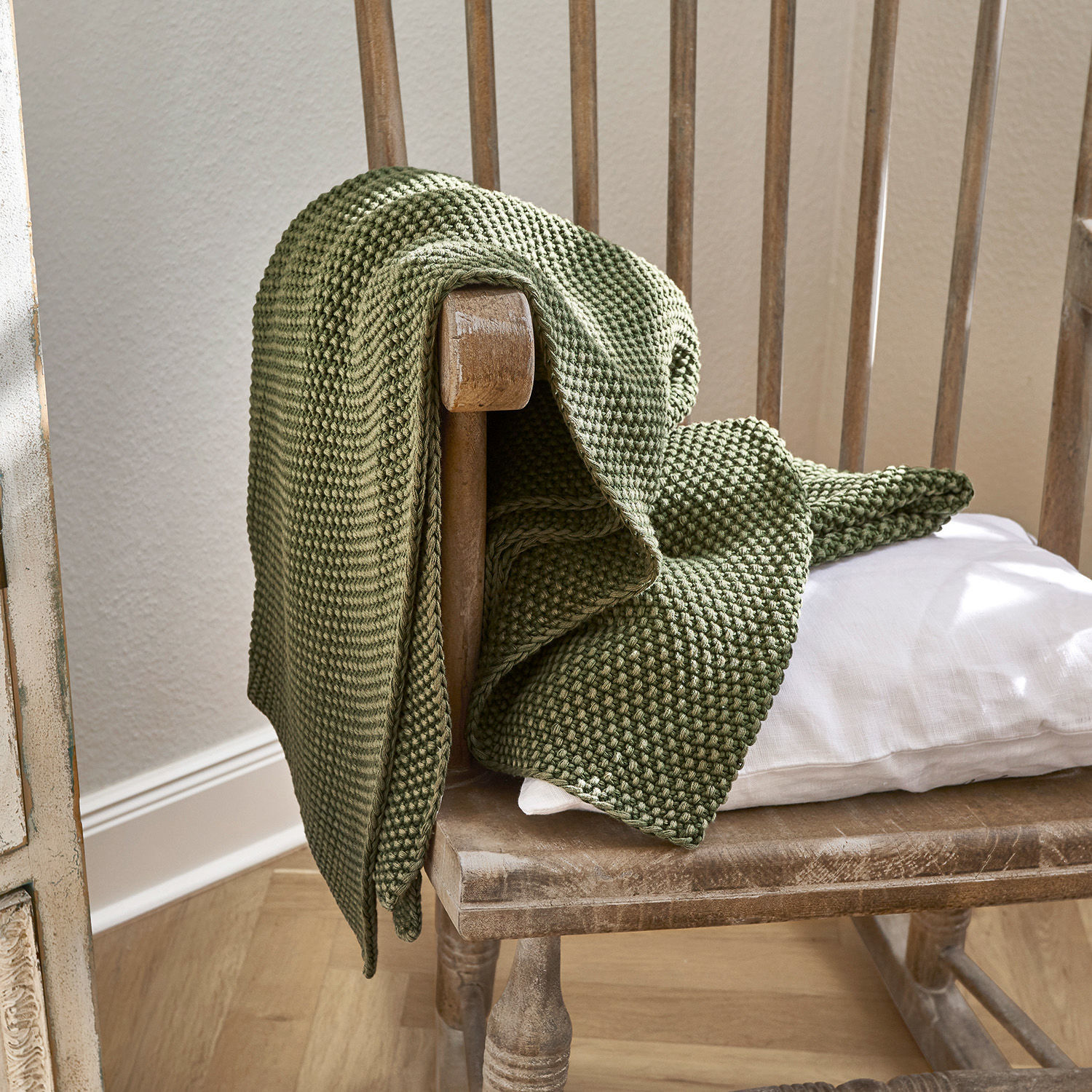Plaid Amberley Plaid vert en coton tricoté posé sur une chaise en bois avec coussin blanc, décoration intérieure cosy