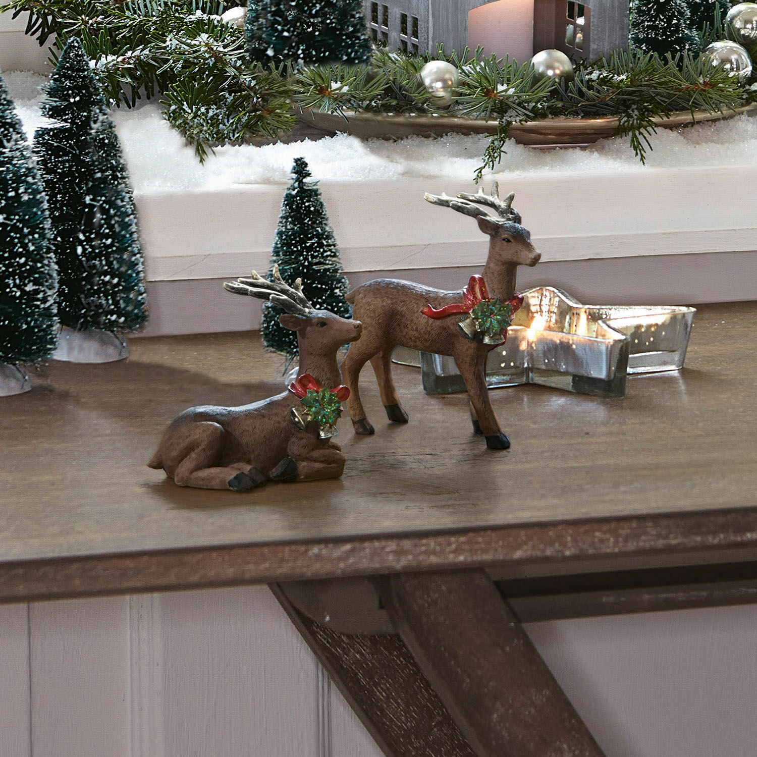 Lot de 2 cerfs Ando Deux figurines de rennes marron avec cloches décoratives et couronnes vertes sur une table en bois