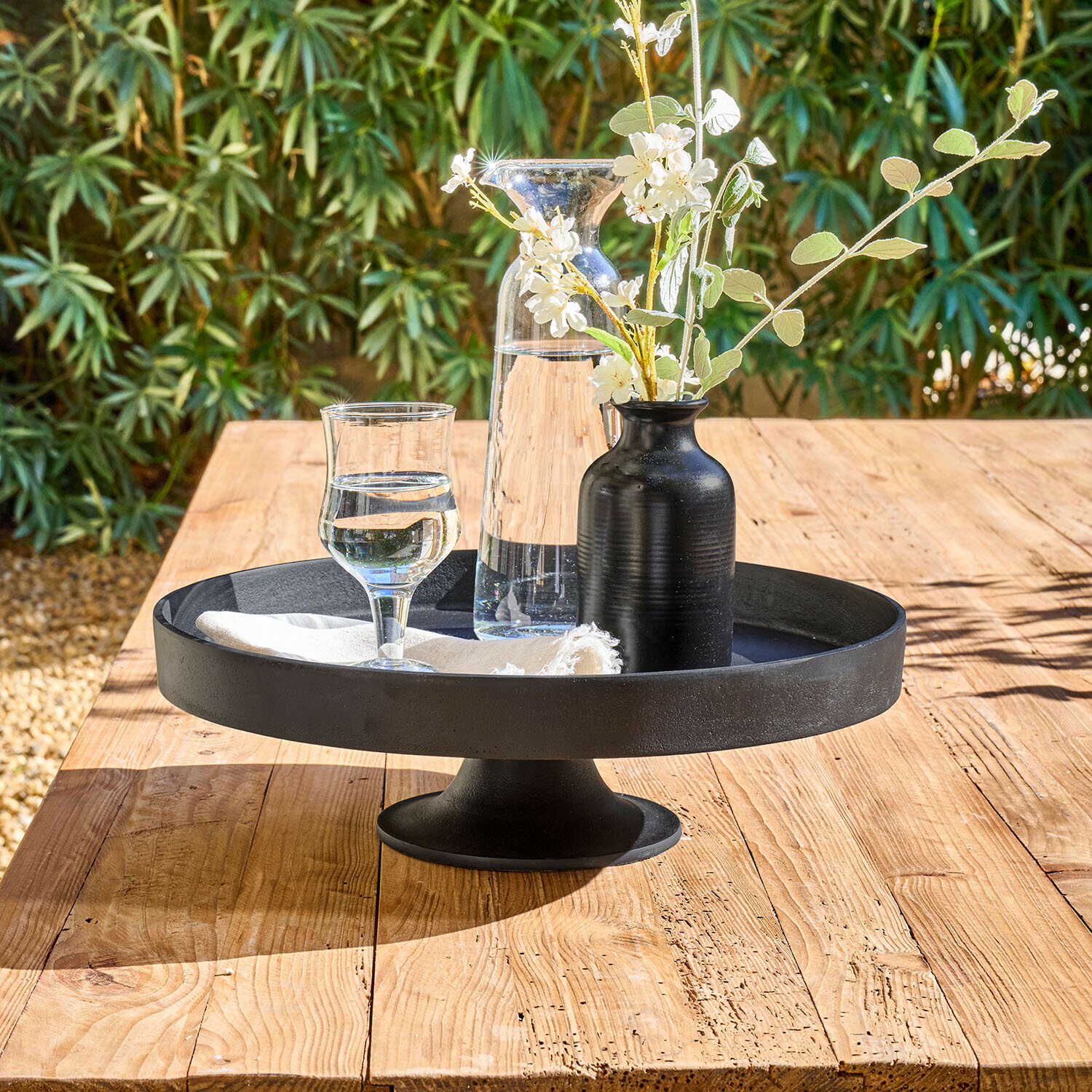 Plateau noir en métal sur pied avec carafe, verre et vase noir fleuri sur table en bois clair