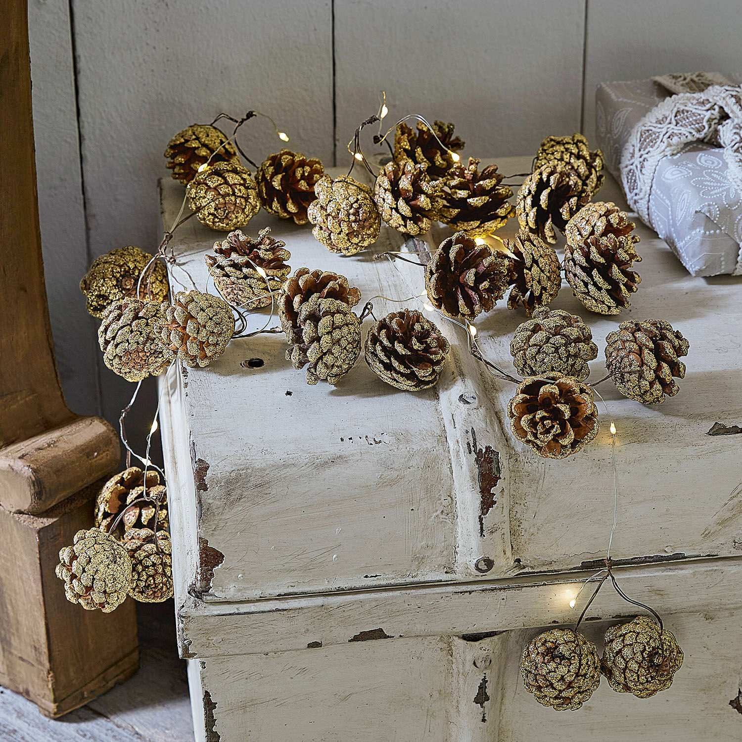 Guirlande lumineuse Bannet Guirlande de pommes de pin naturelles pailletées avec éclairage LED délicat sur meuble en bois blanc