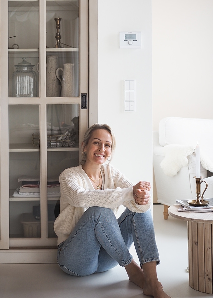 Femme souriante en pull blanc et jean assise par terre dans un intérieur lumineux et chaleureux.