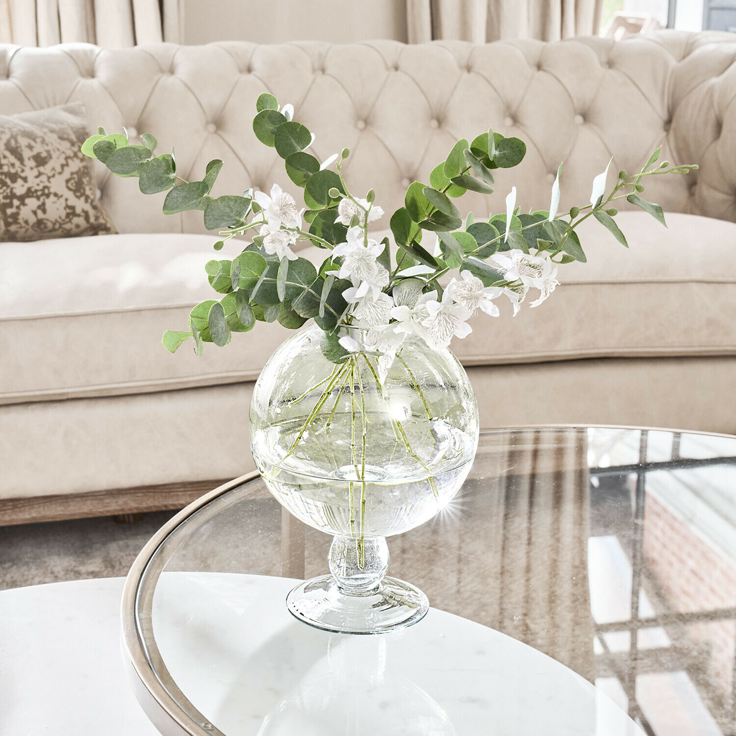 Vase en verre transparent avec feuillage vert et fleurs blanches sur table en verre dans un salon clair
