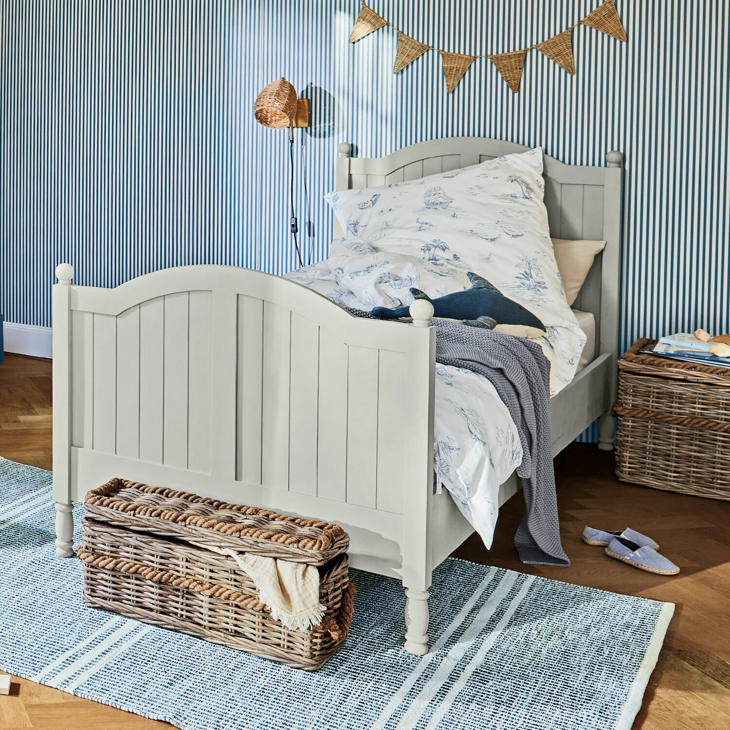 Lit Alarion Lit enfant en bois gris avec literie motif marin et panier en osier sur tapis bleu