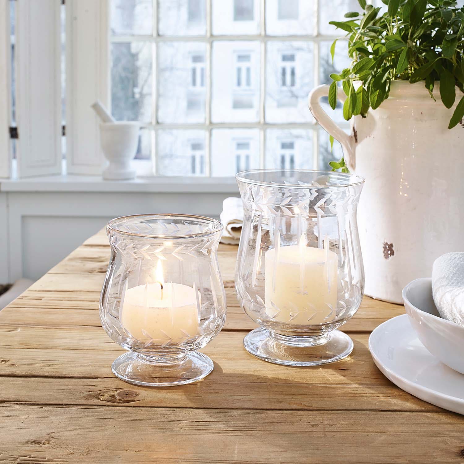 Deux photophores en verre avec bougies blanches sur une table en bois dans une pièce lumineuse