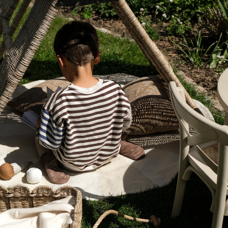 Enfant en pull rayé assis sur un tapis sous une tente en osier dans un jardin, moment de détente
