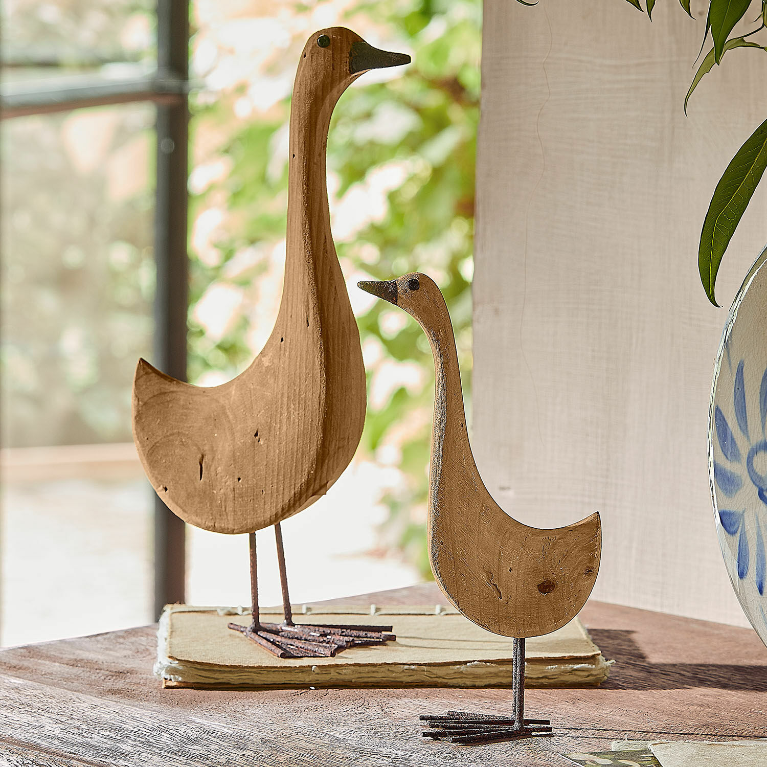 Lot de 2 oies décoratives Amiraz Décorations en bois en forme d'oiseaux couleur naturelle sur pieds métalliques posées sur une table.