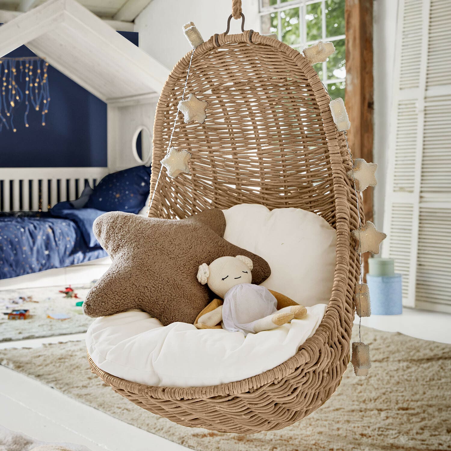 Fauteuil suspendu pour enfant Twinkla Fauteuil suspendu en rotin avec coussin blanc et guirlande lumineuse étoile dans une chambre d'enfant