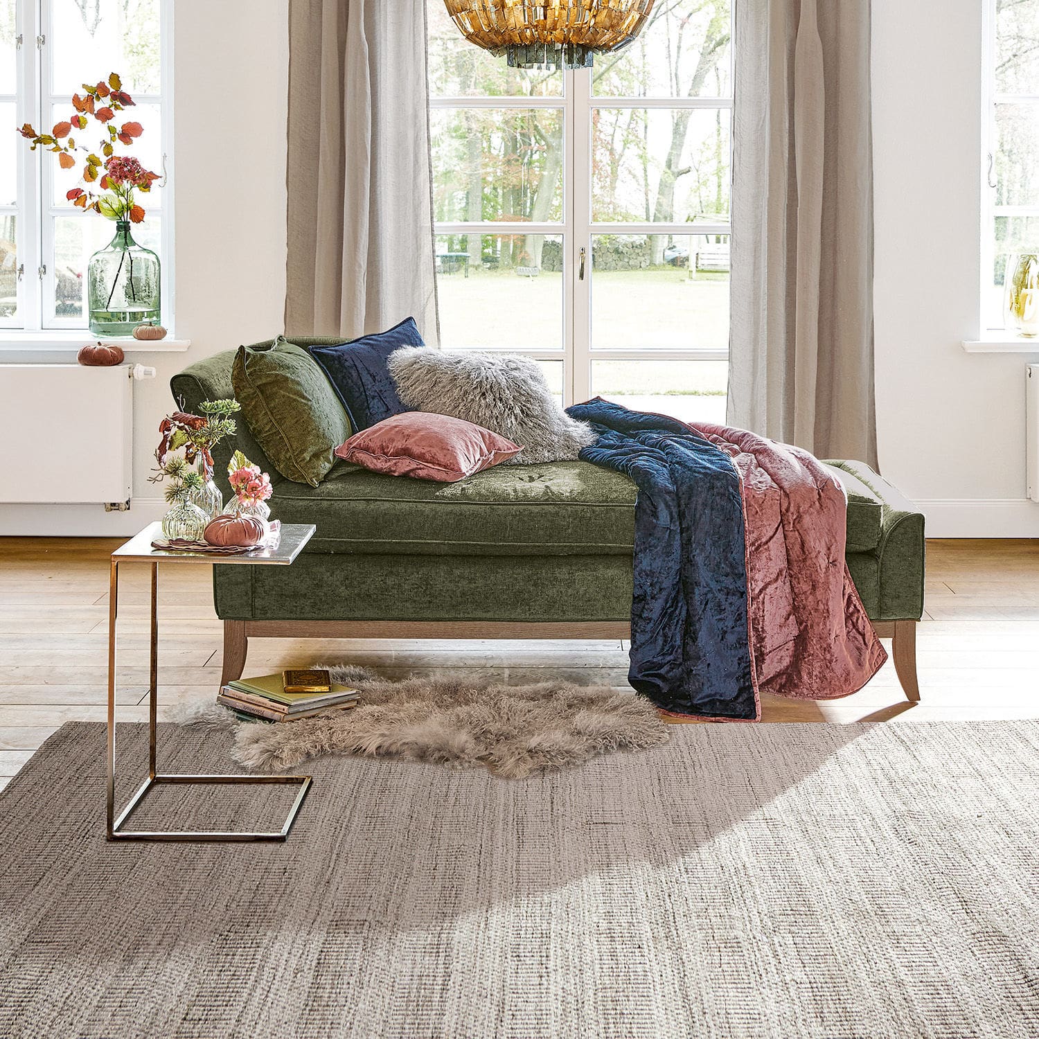 Méridienne Alphonse Chaise longue en velours vert avec coussins et plaid rose et bleu dans un salon lumineux avec grande fenêtre