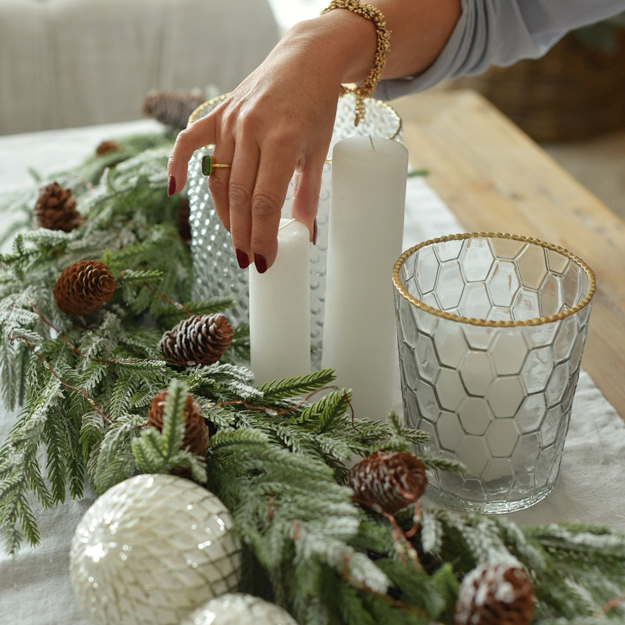 Décoration de Noël avec bougies blanches, photophore en verre à bord doré et branches vertes avec pommes de pin