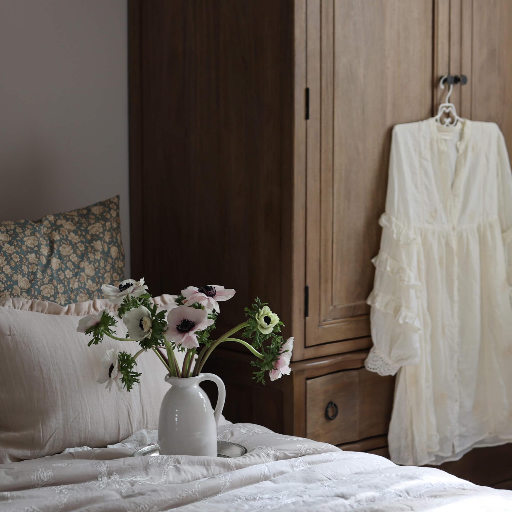 Pichet en céramique blanche avec fleurs sur lit à linge clair devant une armoire en bois