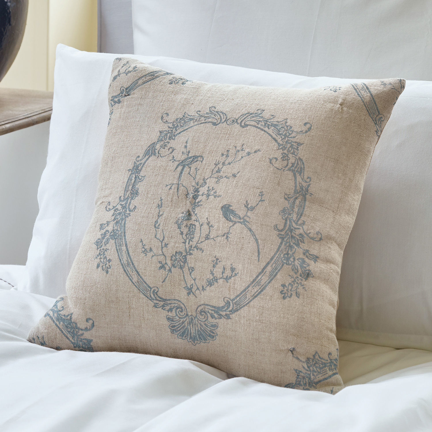 Housse de coussin Decadence Coussin beige en lin avec motif bleu d'oiseaux et fleurs sur lit blanc, décoration intérieure élégante