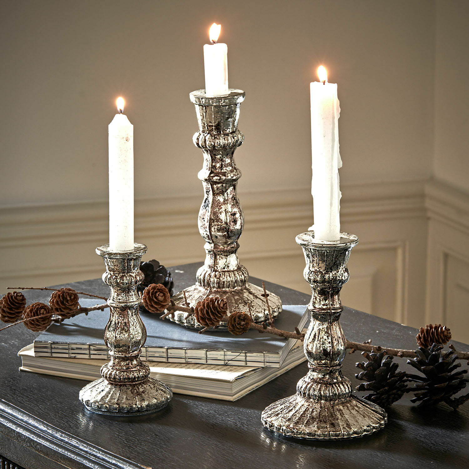 Lot de 3 bougeoirs Favalina Trois chandeliers en métal argenté style vintage avec bougies blanches sur une table sombre décorée de pommes de pin
