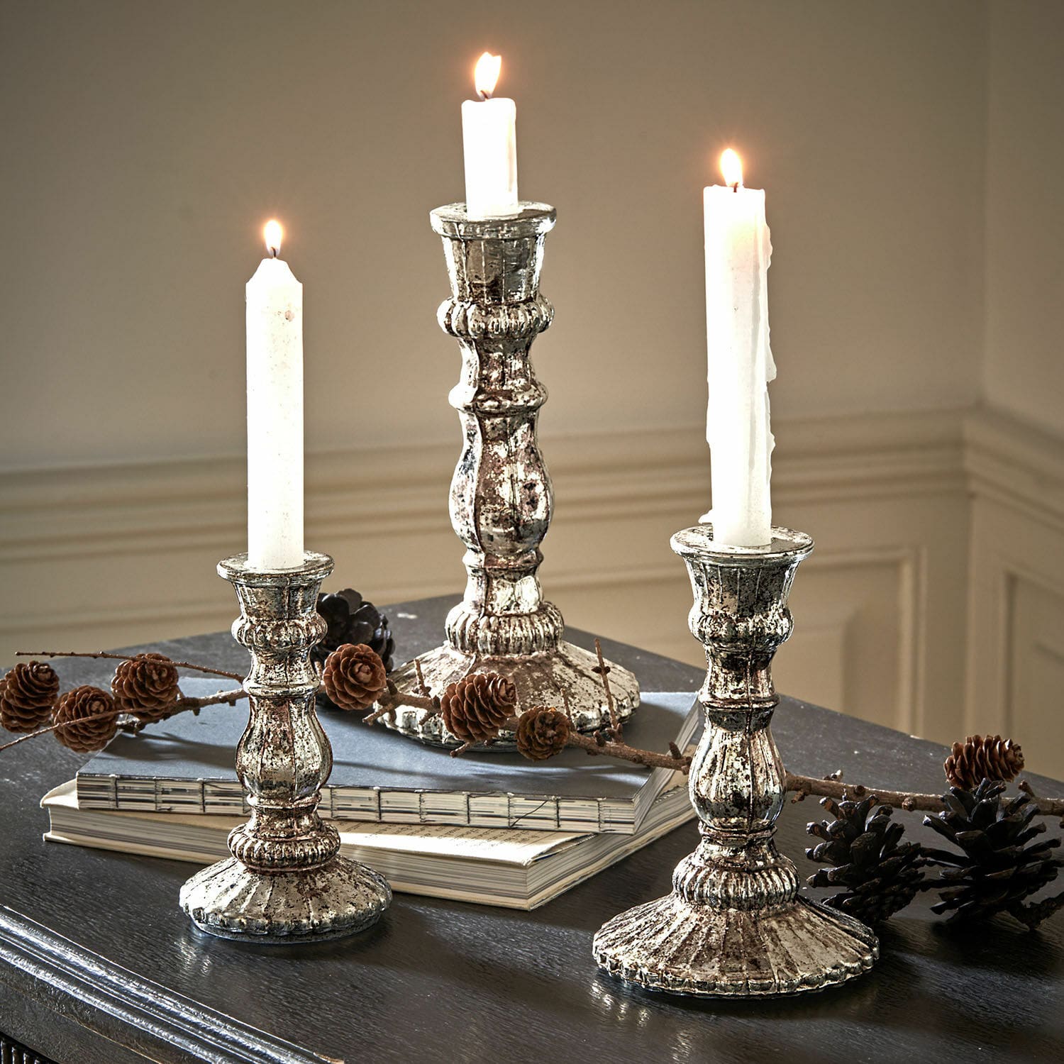 Lot de 3 bougeoirs Favalina Trois chandeliers en métal argenté style vintage avec bougies blanches sur une table sombre décorée de pommes de pin