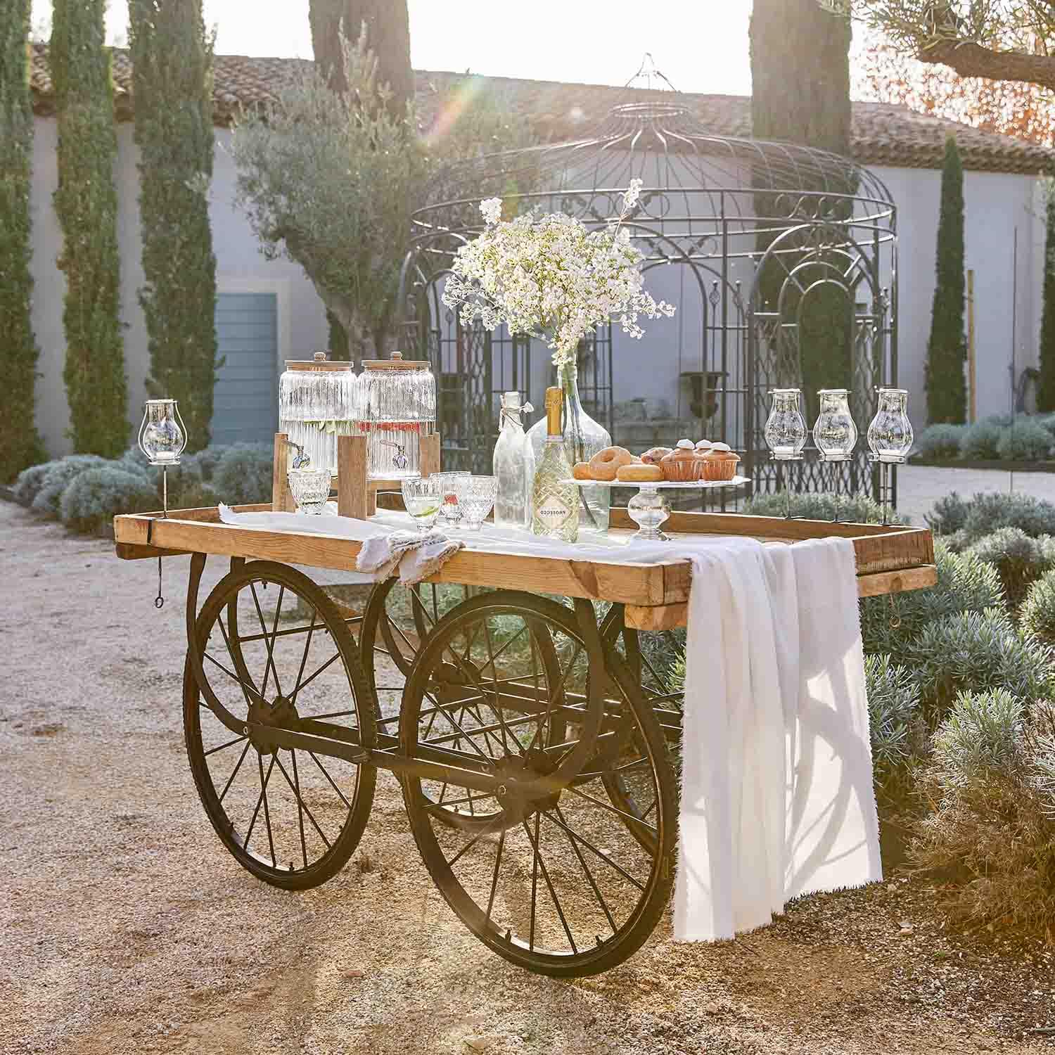 Chariot en bois avec roues en métal décoré de carafes en verre et fleurs dans un jardin ensoleillé