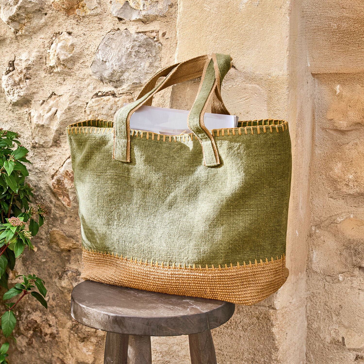 Cabas Yarrowton Sac vert en jute avec finition marron posé sur un tabouret en bois devant un mur en pierre