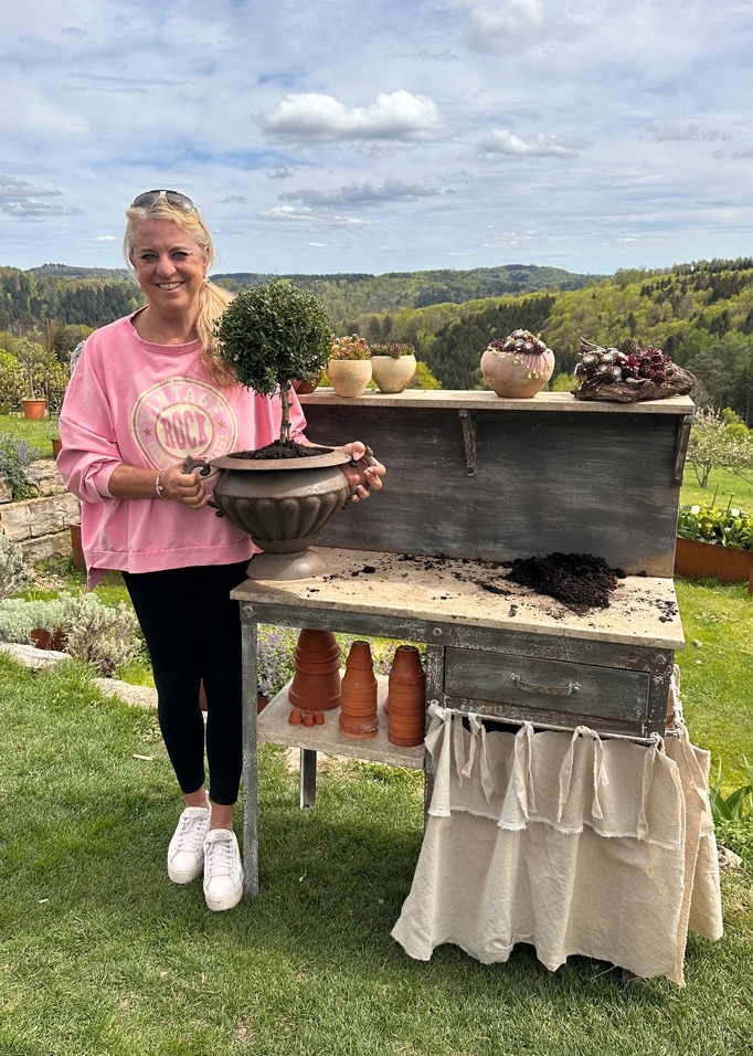 Femme en sweat rose tenant un pot avec un buisson vert près d'une table de jardin en bois avec vue sur la nature