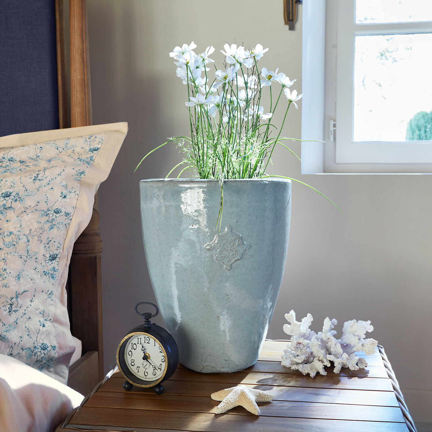 Cache-pot en céramique grise avec fleurs blanches sur table en bois près d'un réveil et décorations marines