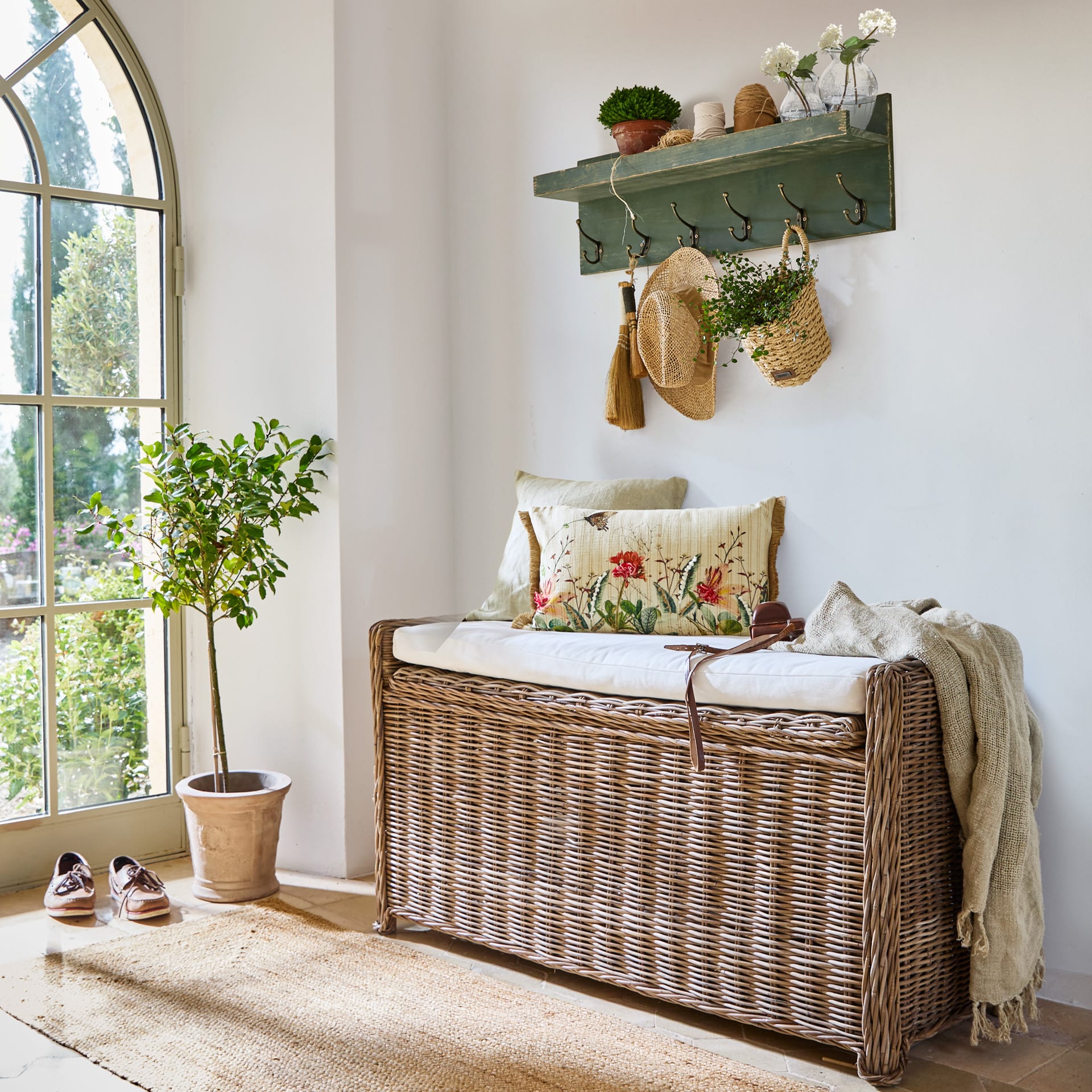 Banc en rotin avec coussin blanc et coussins décoratifs dans un intérieur lumineux avec porte-manteau vert