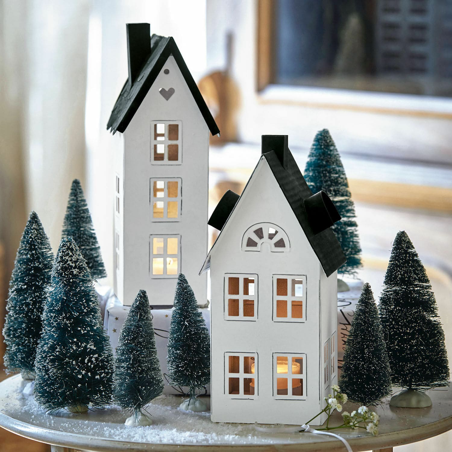 2 lanternes maisonnettes Gunar Maisons décoratives blanches en métal avec toits noirs entourées de sapins artificiels sur une table de Noël
