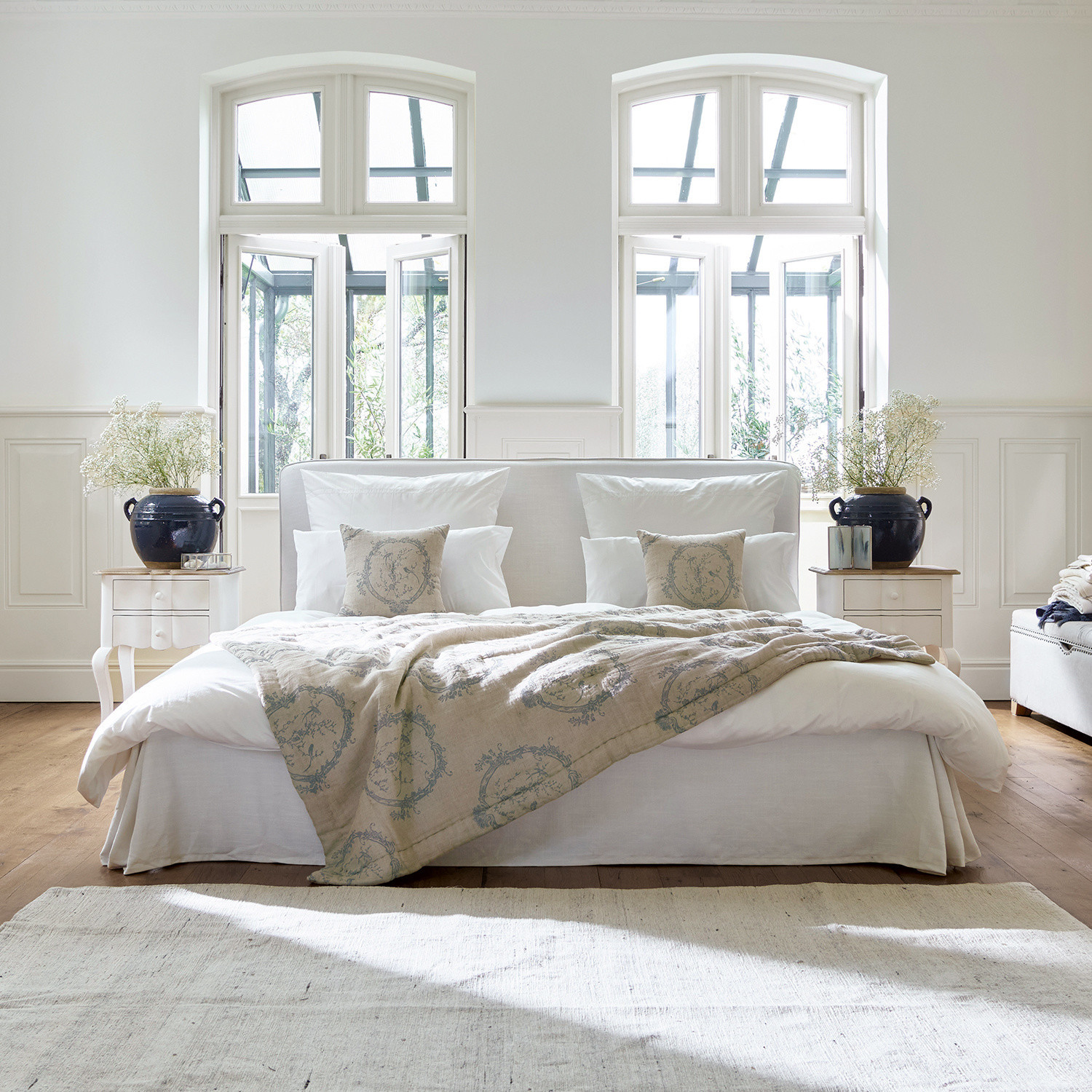 Lit Aubrianne Lit confortable avec revêtement clair et couverture beige à motifs dans une chambre lumineuse avec grandes fenêtres