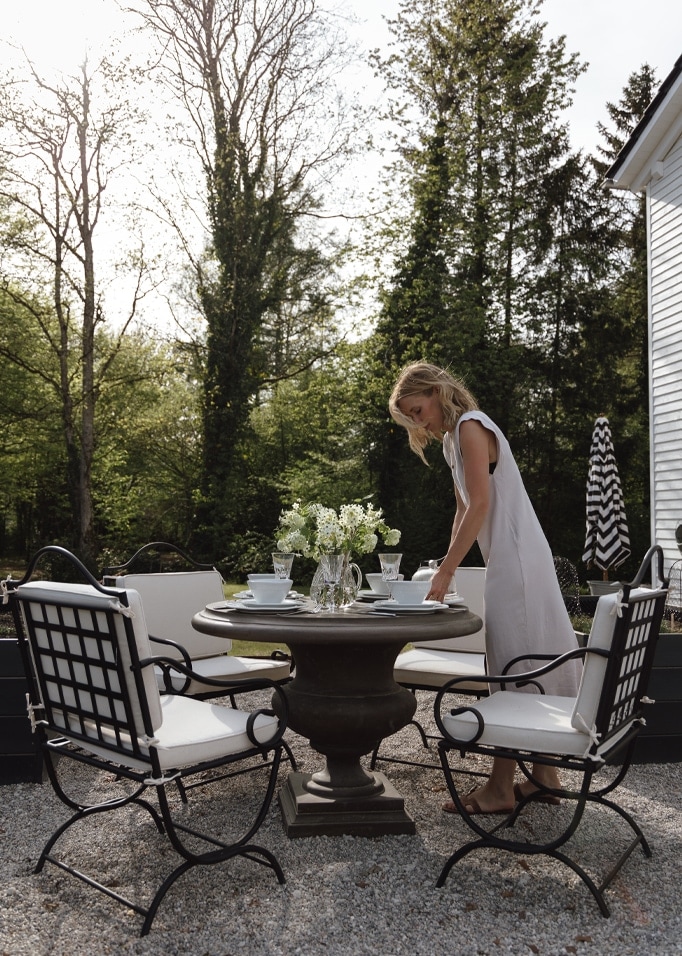 Jardin avec table ronde en pierre et chaises en métal noir avec coussins blancs, femme en robe blanche.