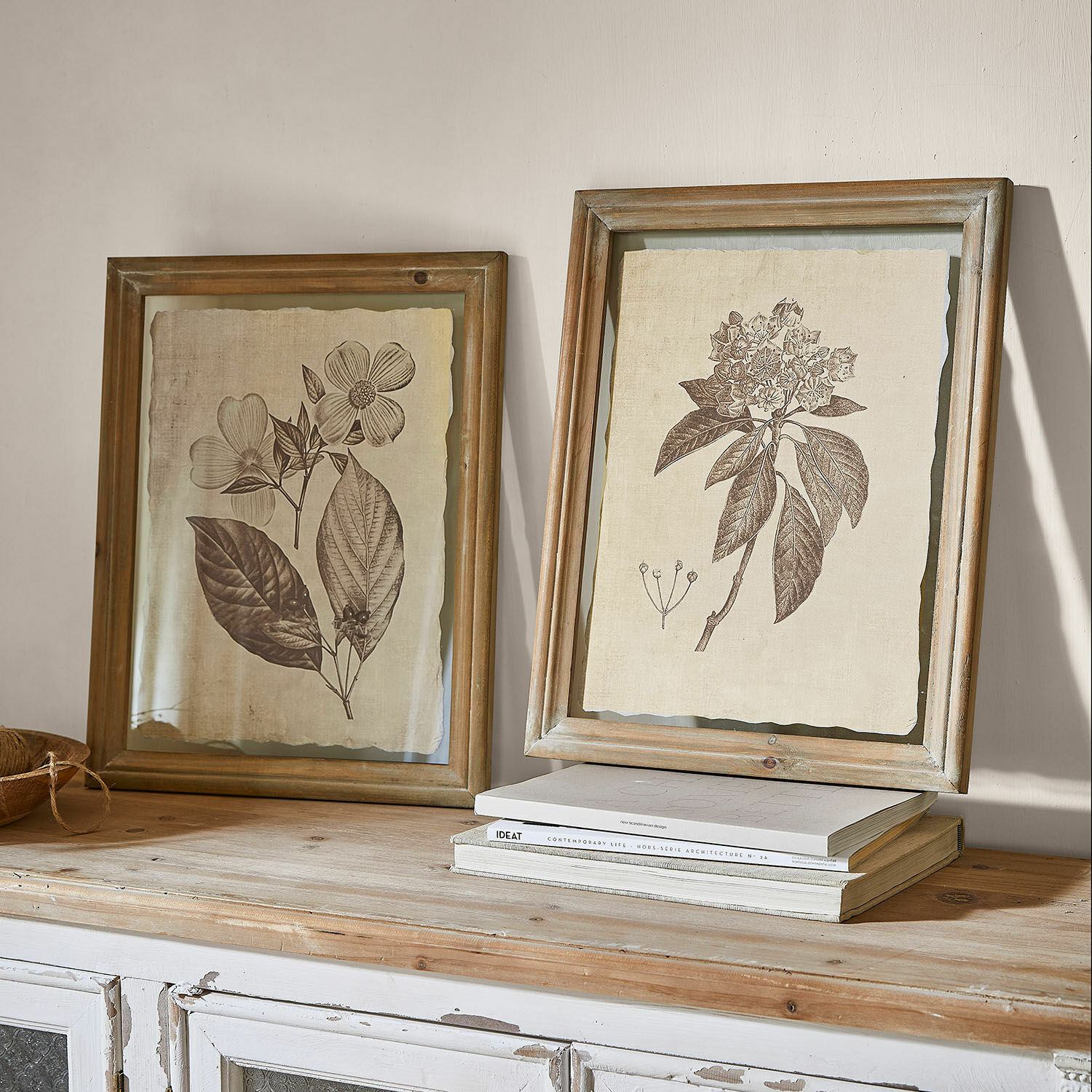 Lot de 2 tableaux Guyra Deux cadres en bois rustiques avec dessins botaniques de fleurs sur fond beige posés sur un meuble en bois