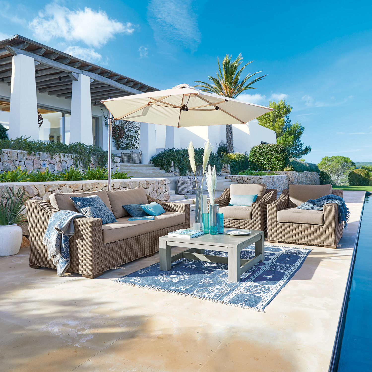 Salon de jardin Chestingdale Ensemble de mobilier de jardin en rotin beige avec parasol et tapis bleu sur terrasse ensoleillée