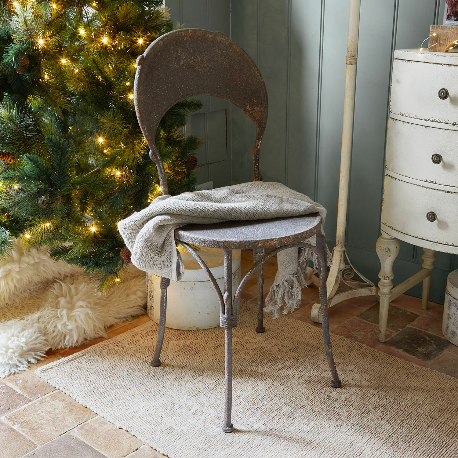 Chaise Delivora Chaise en métal rouillé avec plaid gris posée près d'un sapin de Noël illuminé et tapis beige