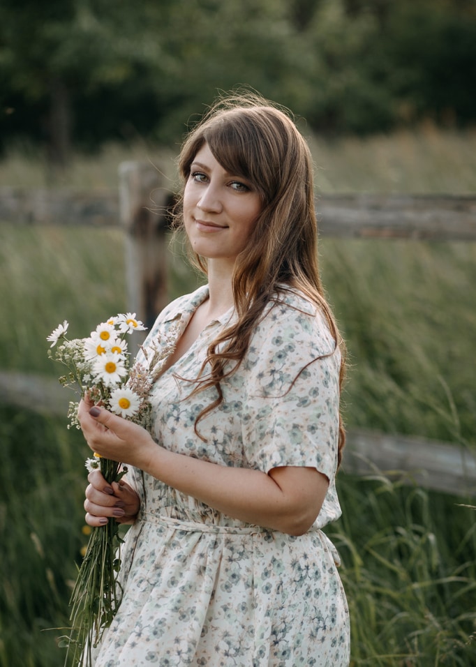 Femme en robe fleurie tenant un bouquet de marguerites devant un champ vert et une clôture en bois