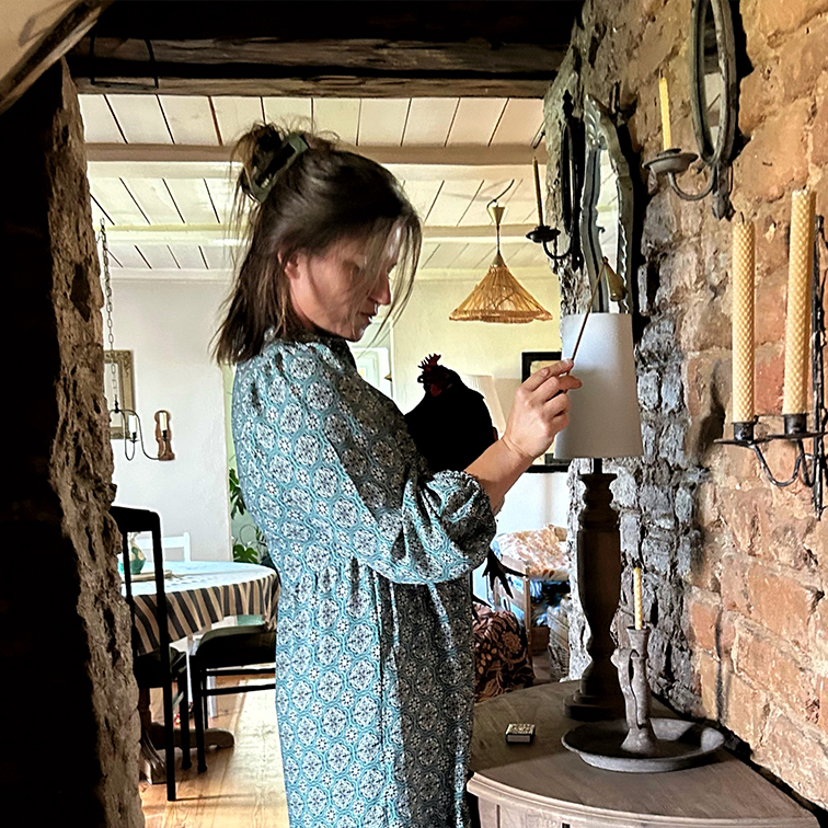 Femme en robe bleue à motifs allumant une lampe dans un intérieur rustique avec mur en briques et bougies