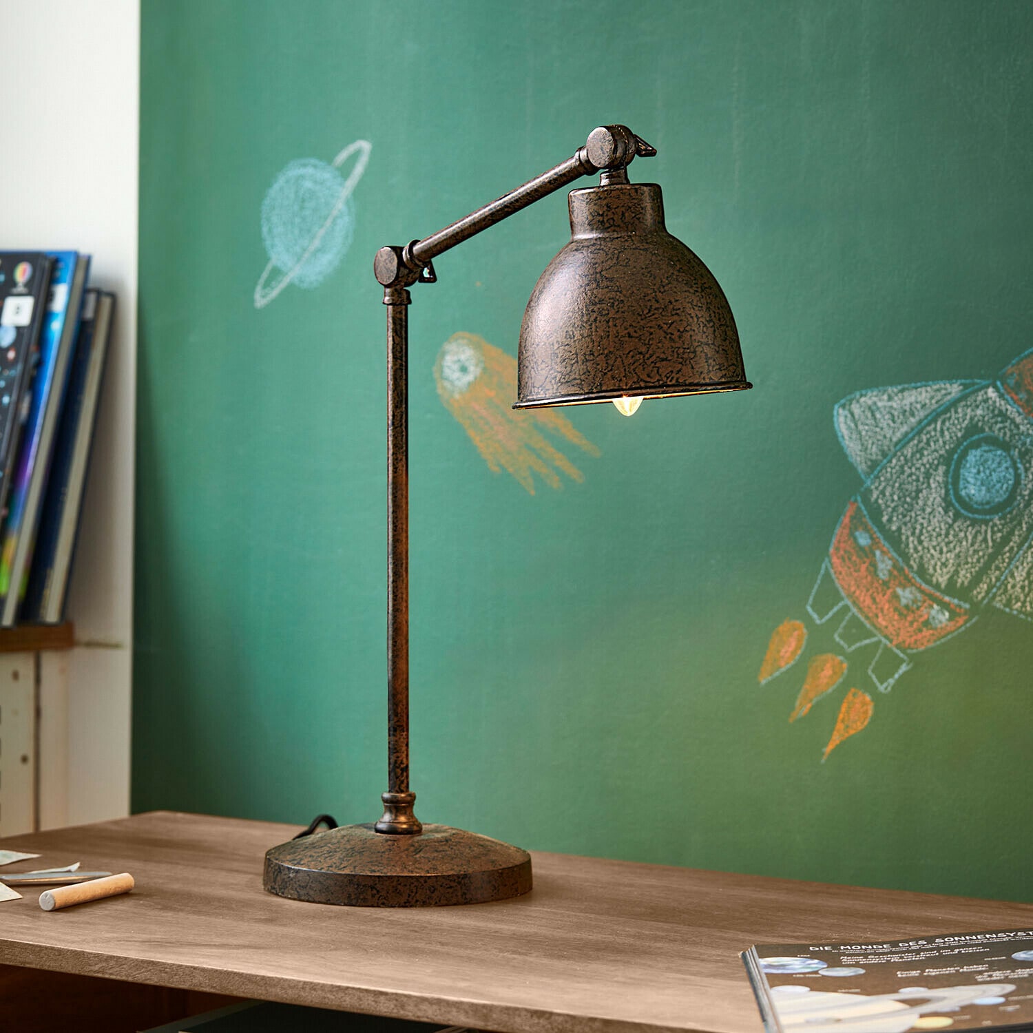 Lampe de bureau Cheeklee Lampe de bureau en métal effet rouille sur un bureau en bois devant un tableau vert avec dessins d'espace
