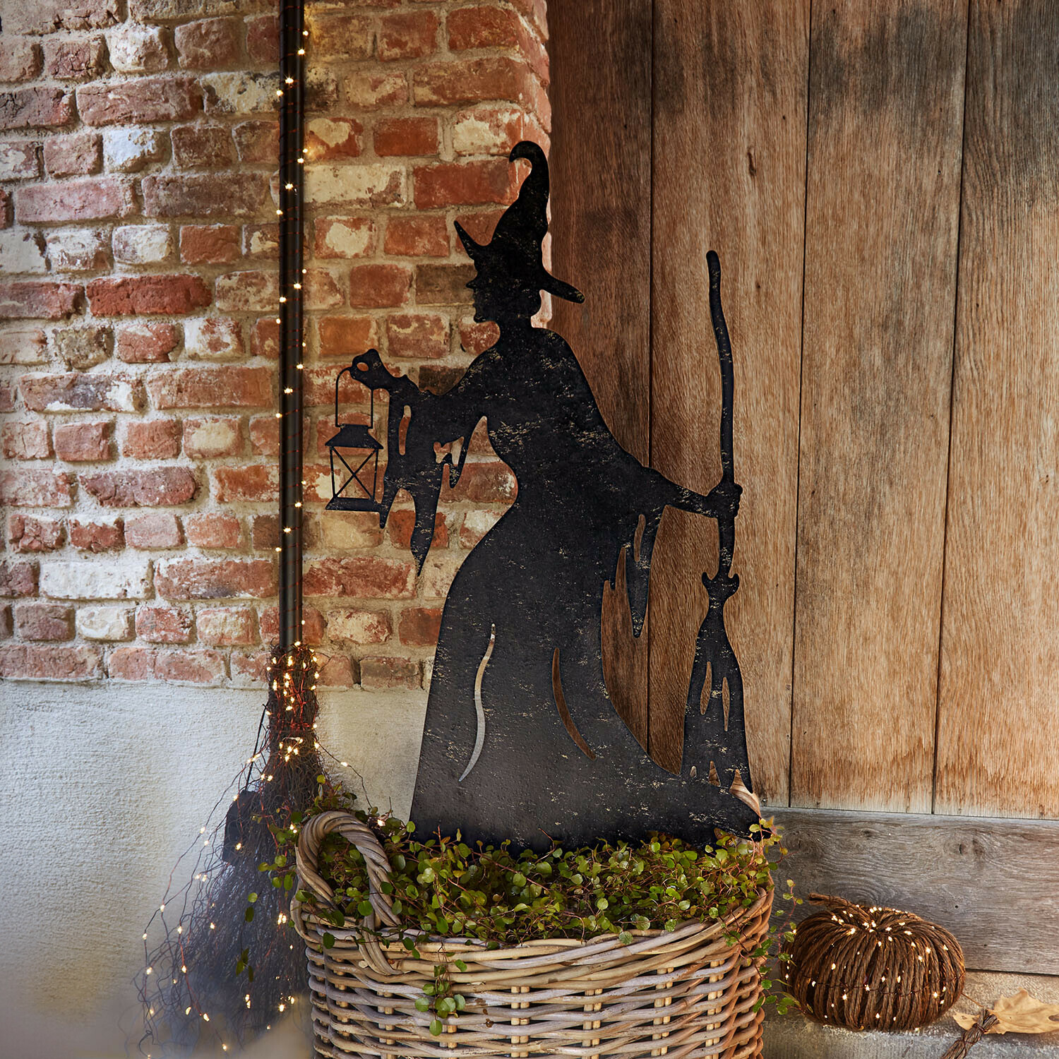 Pique de jardin Talmora Décoration en métal noir silhouette de sorcière avec lanterne et balai près d'un panier en osier