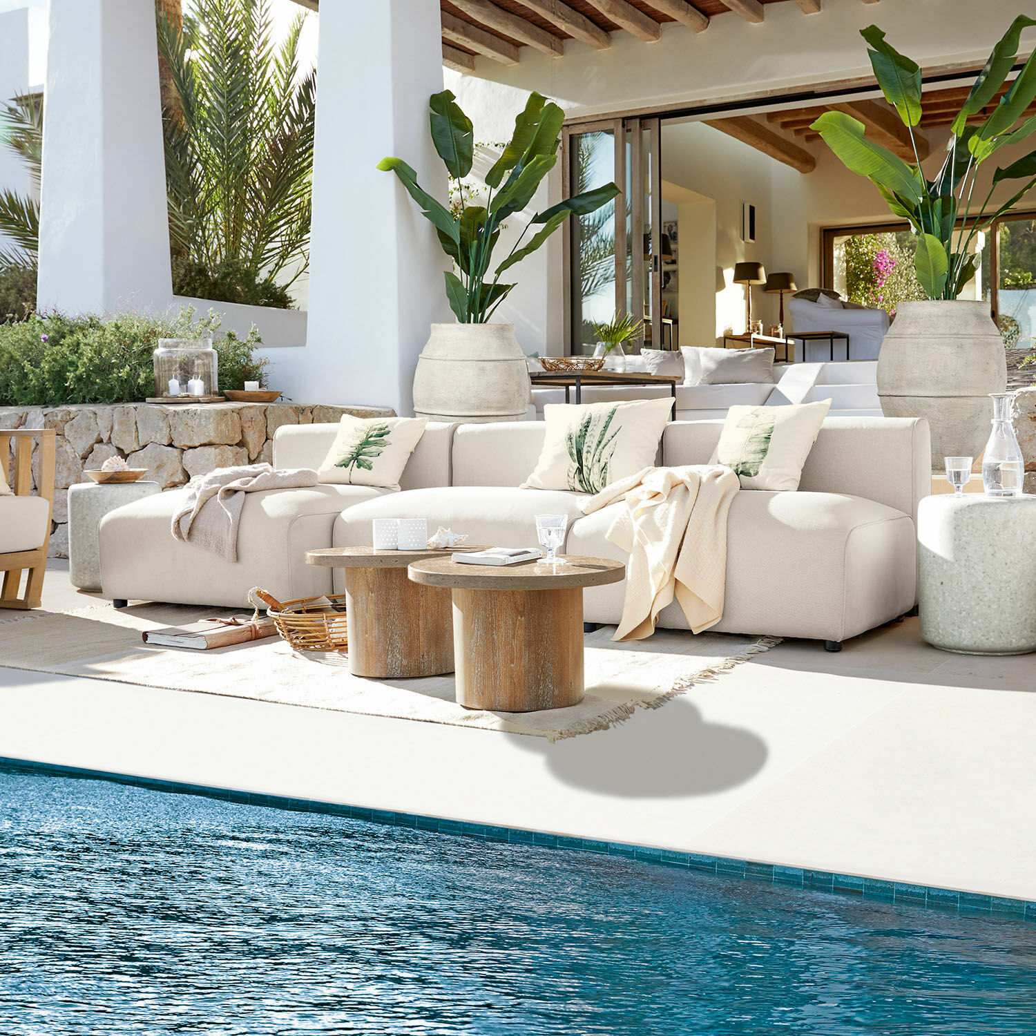 Salon de jardin Vézère Canapé beige en tissu sur terrasse au bord de la piscine avec tables basses en bois et coussins décoratifs