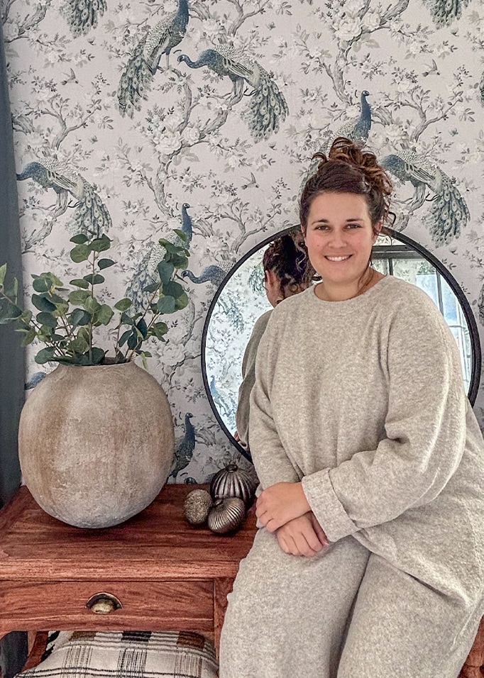 Femme en pull gris assise près d'une table en bois avec un grand vase en céramique et un miroir devant un papier peint à motif paon