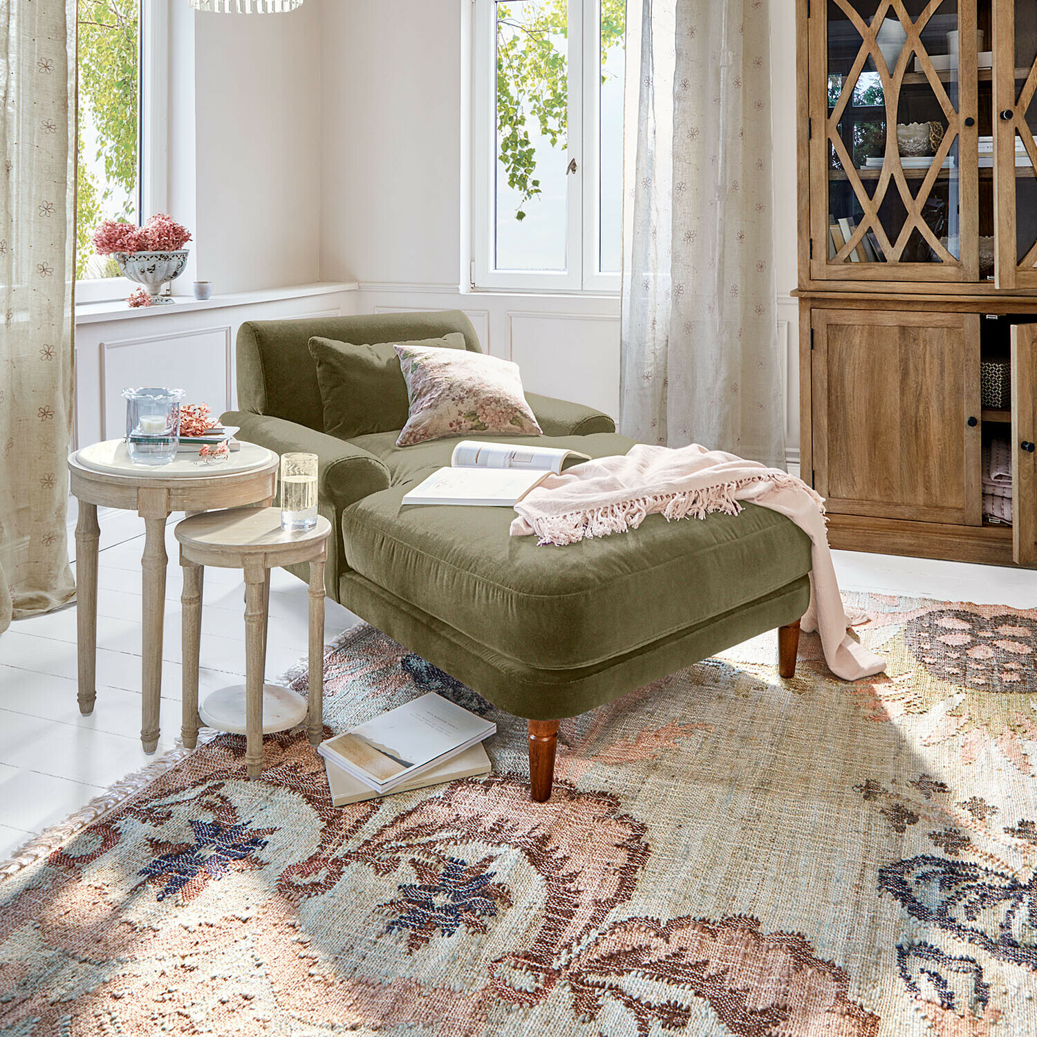 Chaise longue en velours vert olive avec coussins et plaid rose dans un salon lumineux avec meubles en bois