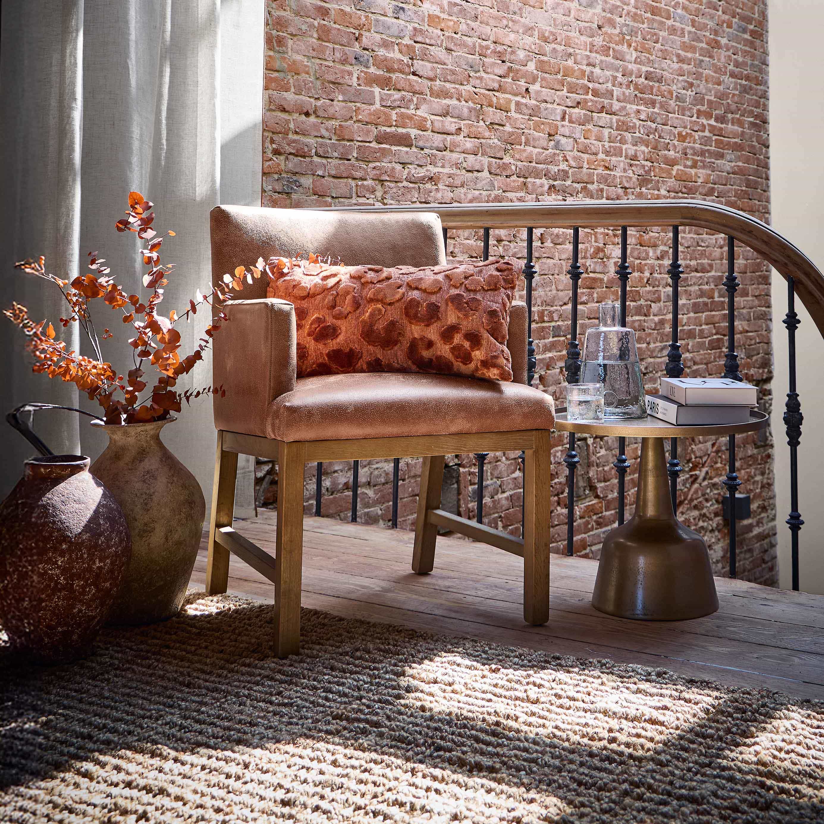 Fauteuil beige avec base en bois et coussin orange devant un mur en briques et des rideaux