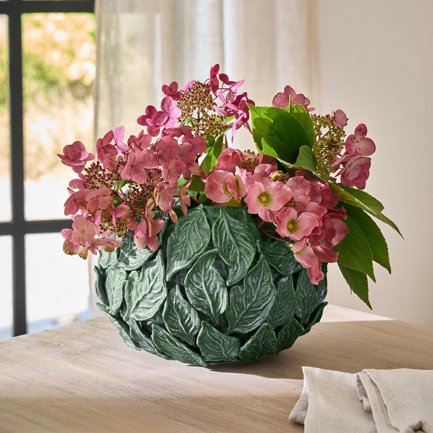 Vase décoratif vert en forme de feuilles avec fleurs roses sur table en bois dans une pièce lumineuse