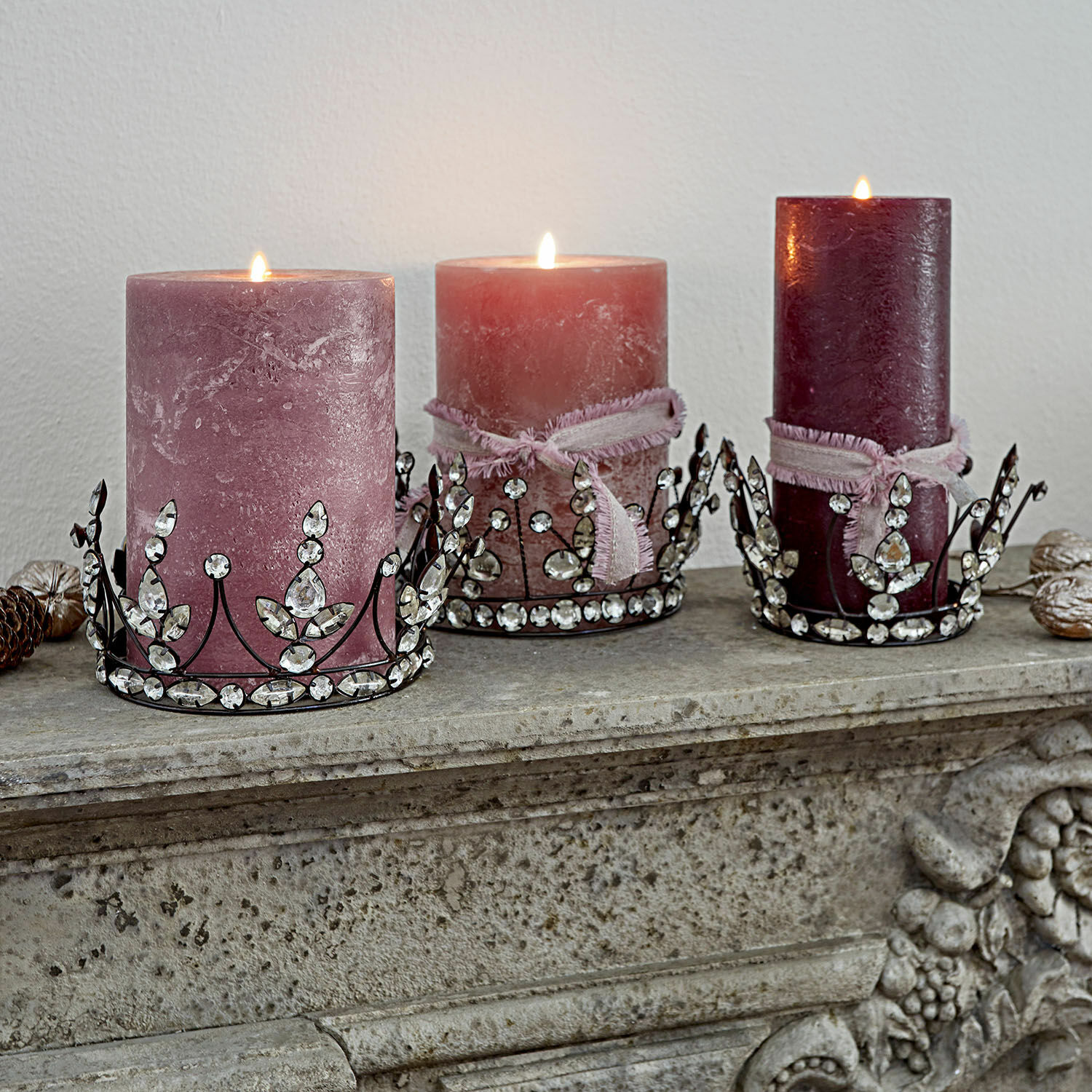 Lot de 3 couronnes décoratives Bigas Trois bougies roses décoratives avec supports métalliques ornés de cristaux sur une cheminée en pierre