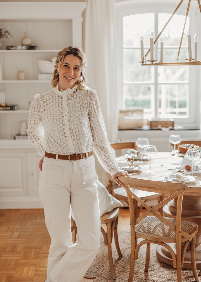 Femme en blouse blanche en dentelle et pantalon clair près d'une table en bois dans une pièce lumineuse.