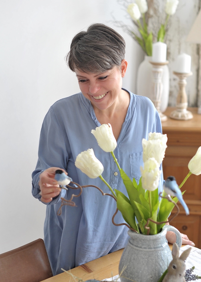 Femme en chemise bleue décorant une table avec un vase en céramique grise, tulipes blanches et oiseaux décoratifs