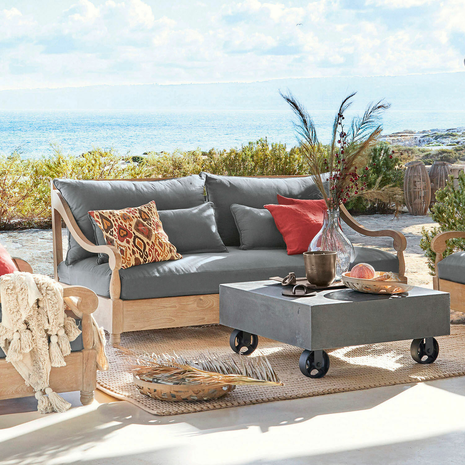 Canapé Montcassin Canapé d'extérieur en bois clair avec coussins gris et coussins décoratifs colorés sur terrasse face à la mer