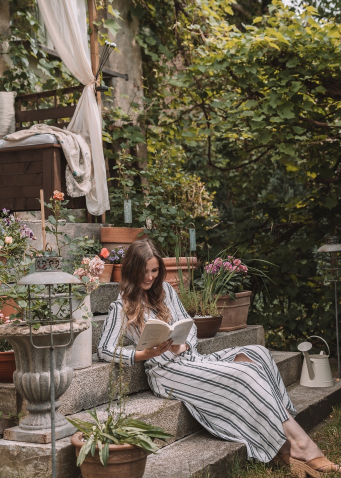 Femme en robe rayée lisant un livre sur des marches en pierre entourée de pots de fleurs dans un jardin
