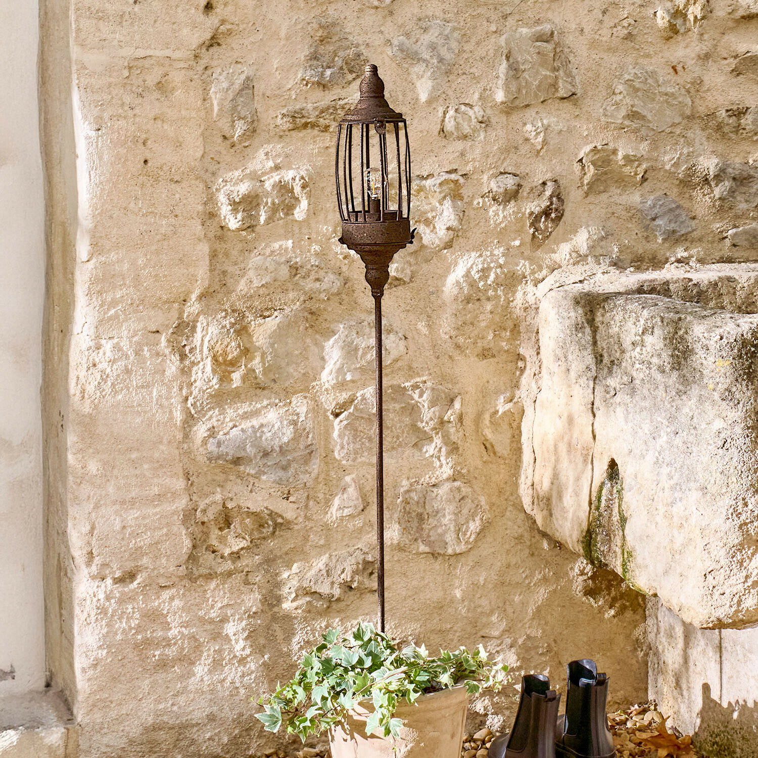 Lampe de jardin en métal rouillé sur pied long devant un mur en pierre rustique