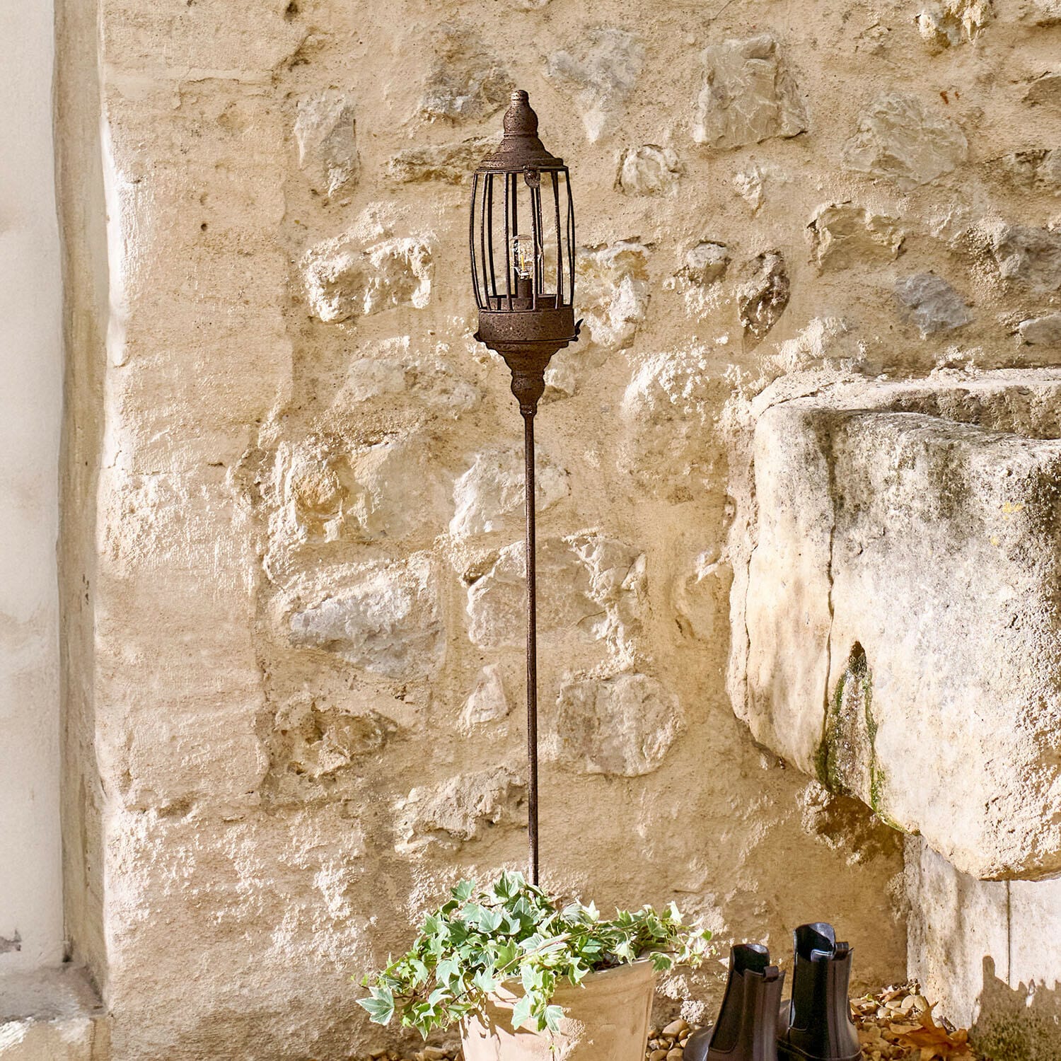 Lanterne LED rechargeable Lagia Lampe de jardin en métal rouillé sur pied long devant un mur en pierre rustique