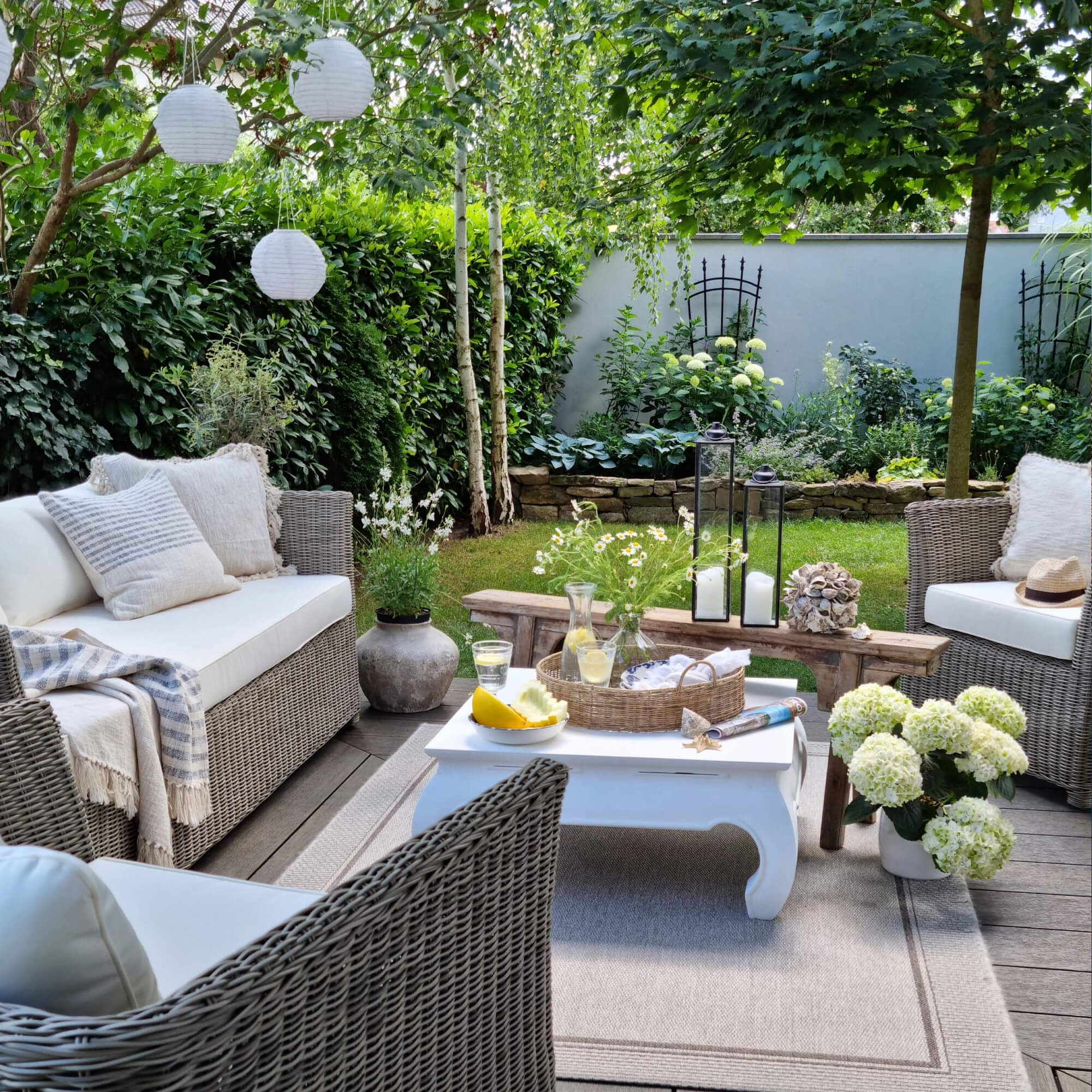 Coin jardin confortable avec mobilier en rotin gris et coussins blancs, décorations et plantes vertes.